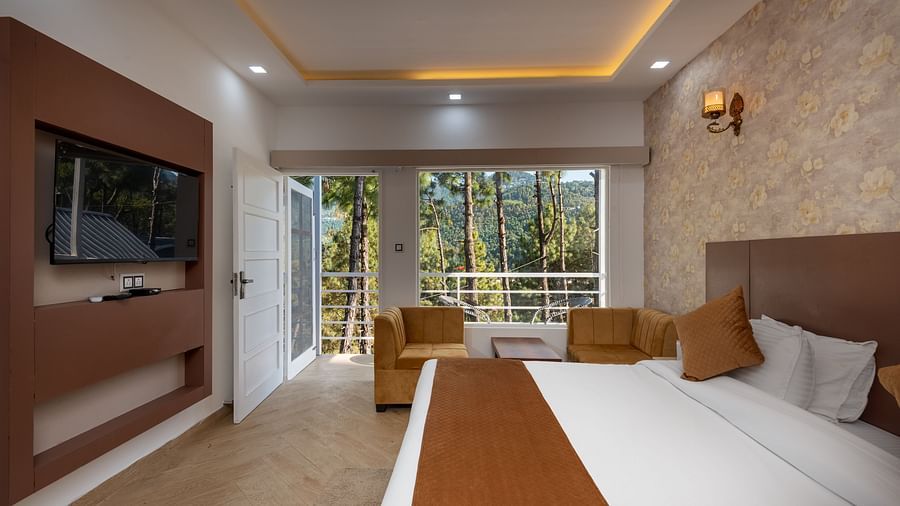 Bright hotel room with a brown and beige theme, featuring a double bed, flat-screen TV, cosy seating area, and large windows overlooking a forested view - DLS Nature Trinket Resort, Dalhousie.
