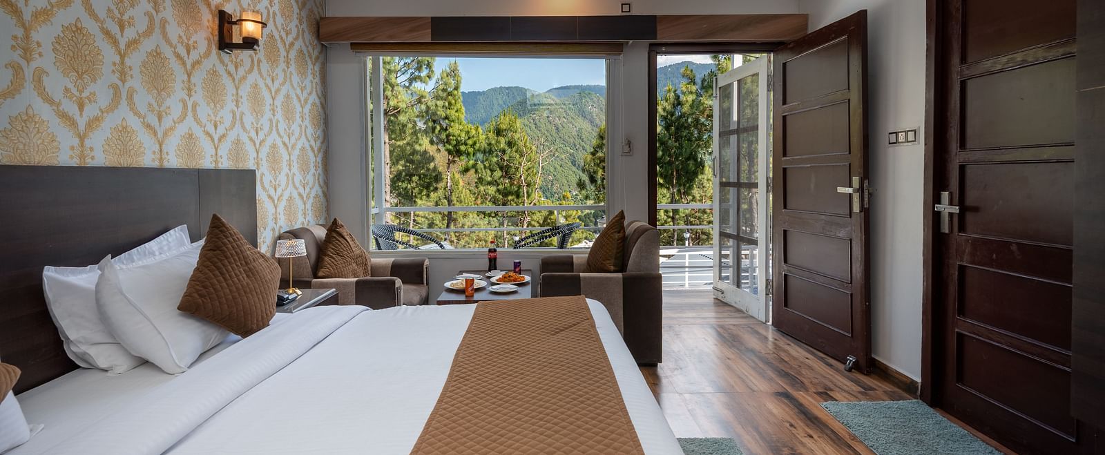  Stylish hotel room featuring a neatly made bed with brown accents, two wooden doors, a sitting area near a large window, and a balcony that opens to a scenic view of trees and distant hills - DLS Nature Trinket Resort, Dalhousie.