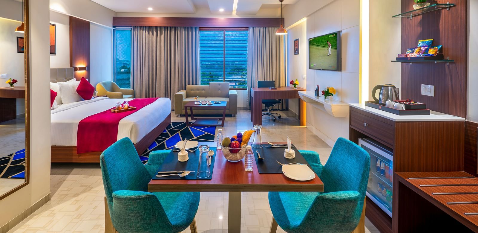 A hotel suite with a bed featuring blue and white bedding, a dining table with blue chairs, and a view of the outside.