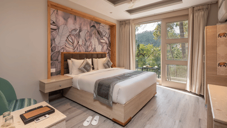 Bright Deluxe Room at DLS Whispering Winds, Kasauli featuring a king size bed, mountain-inspired headboard artwork, and large windows overlooking greenery.