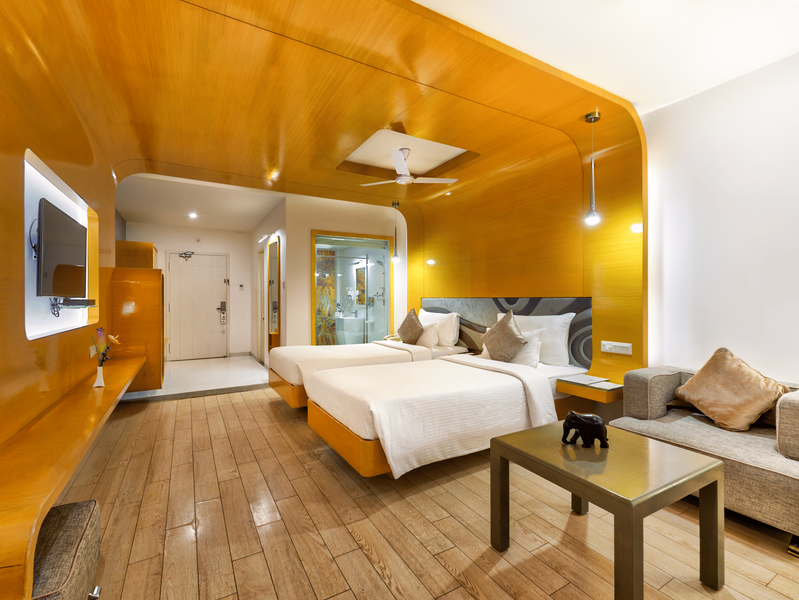 A room with twin beds, golden wall panel and wooden furniture featuring a seating space for a comfortable stay - Mahabalipuram's Grande Bay Resort & Spa.