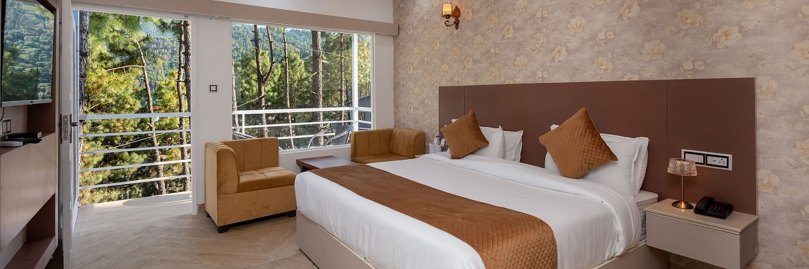 Spacious hotel room with a large double bed, neutral-toned décor, ceiling fan, wall-mounted lights, beige armchair, TV, and a balcony view of lush green trees - DLS Nature Trinket Resort, Dalhousie.