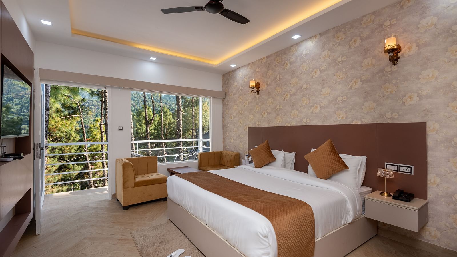 Spacious hotel room with a large double bed, neutral-toned décor, ceiling fan, wall-mounted lights, beige armchair, TV, and a balcony view of lush green trees - DLS Nature Trinket Resort, Dalhousie.