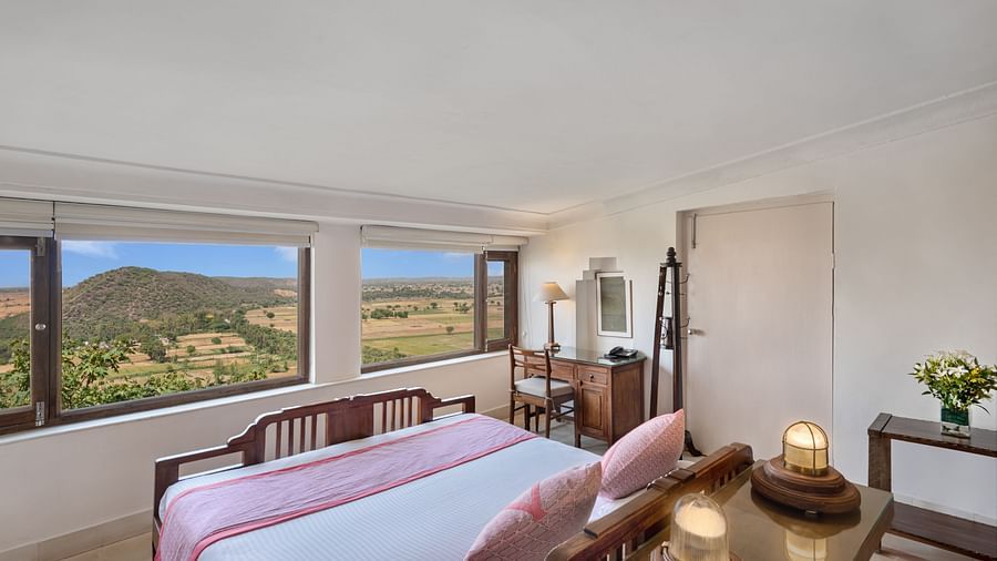 A room with a daybed, a desk, a mirror, a small table with flowers, and large windows with a scenic view in Tijara Fort-Palace - 19th Century, Alwar.
