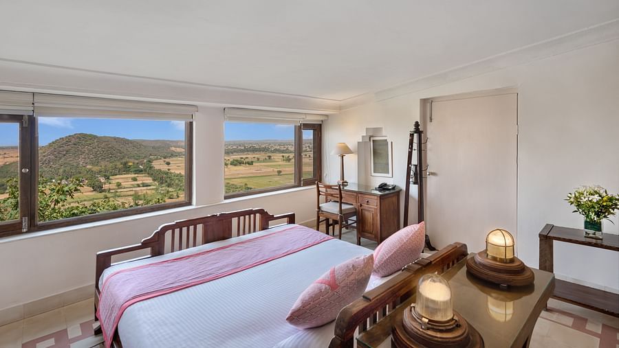 A room with a daybed, a desk, a mirror, a small table with flowers, and large windows with a scenic view in Tijara Fort-Palace - 19th Century, Alwar.