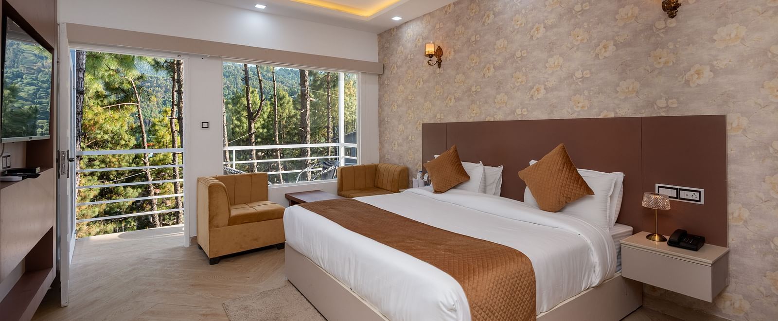 Spacious hotel room with a large double bed, neutral-toned décor, ceiling fan, wall-mounted lights, beige armchair, TV, and a balcony view of lush green trees - DLS Nature Trinket Resort, Dalhousie.
