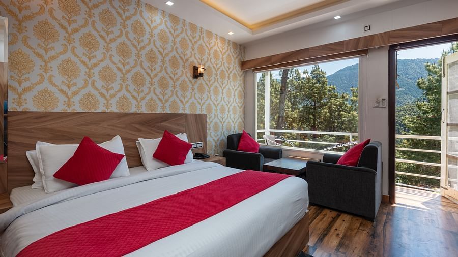  Elegant hotel room with a large bed featuring red and white bedding, stylish wallpaper, a cosy seating area, and a balcony with scenic mountain and forest views - DLS Nature Trinket Resort, Dalhousie.