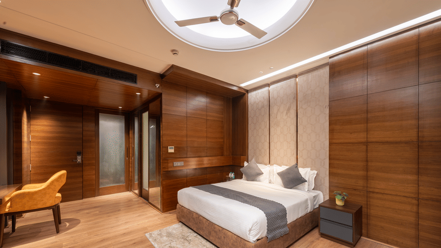 A bedroom with a king size bed, side tables, chair, wardrobe, and ceiling fan inside at DLS Hotels Amritsar.