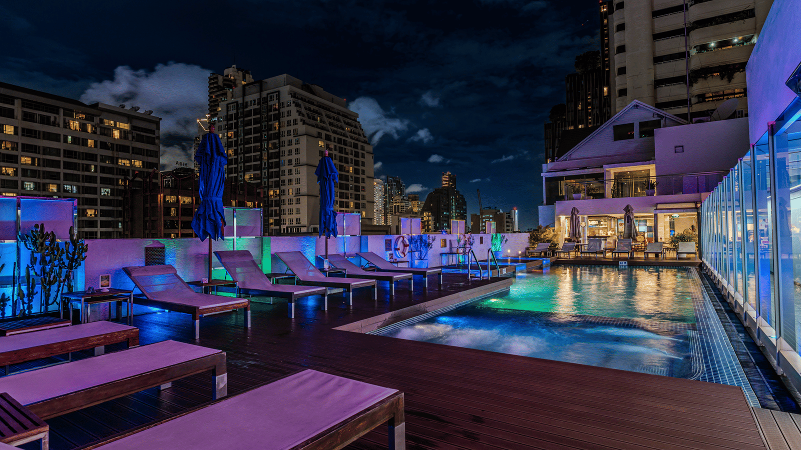 An overview of the rooftop swimming pool with loungers in front of it and high rise buildings next to it inside Night Hotel Bangkok, a hotel near Nana BTS Bangkok.