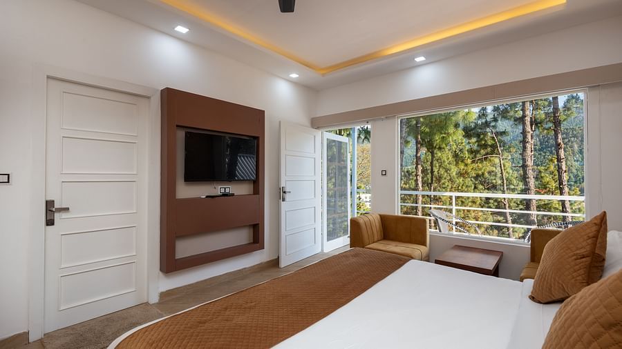 Modern hotel room with a double bed featuring brown accents, a wall-mounted flat-screen television, comfortable armchairs, and large windows overlooking a forested view - DLS Nature Trinket Resort, Dalhousie.