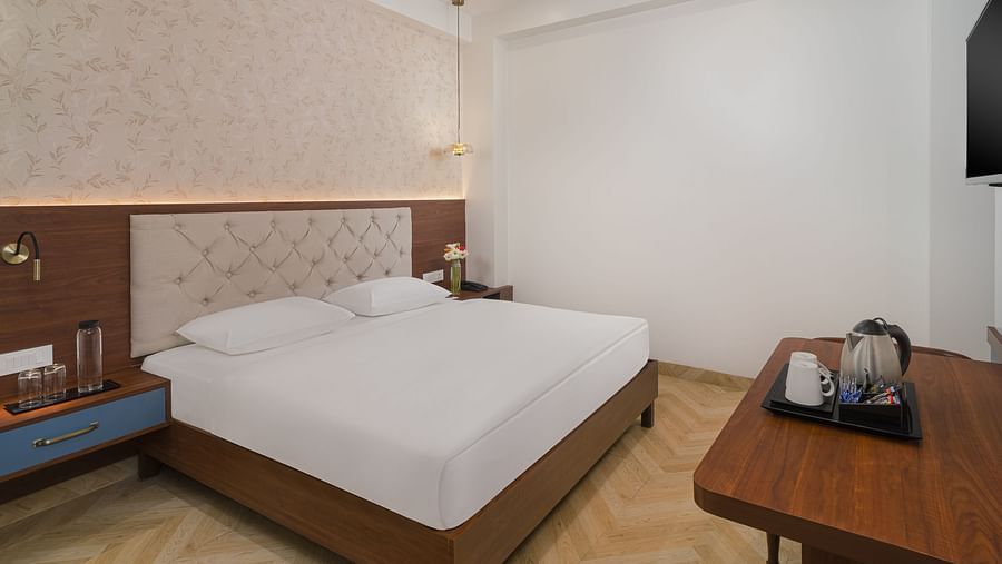 Side view of a spacious room featuring a large bed with a cushioned headboard, wooden bedside tables on either side, and inviting lighting | Hotel Ganga Maiya