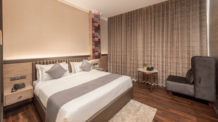 A bedroom with a king size bed, size tables, chair, curtains, and table inside at DLS Hotels Amritsar.