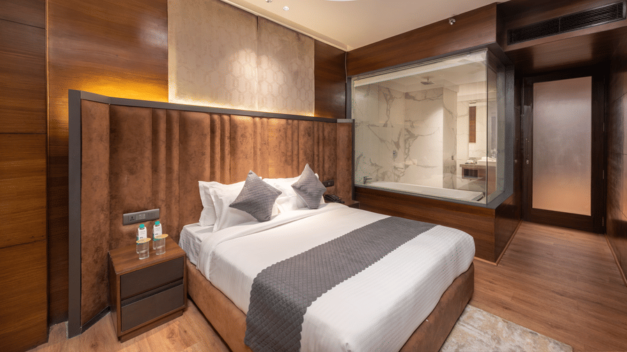 A room with a king size bed, side table, ceiling fan, and a glass partition leading to another area at DLS Hotels Amritsar.