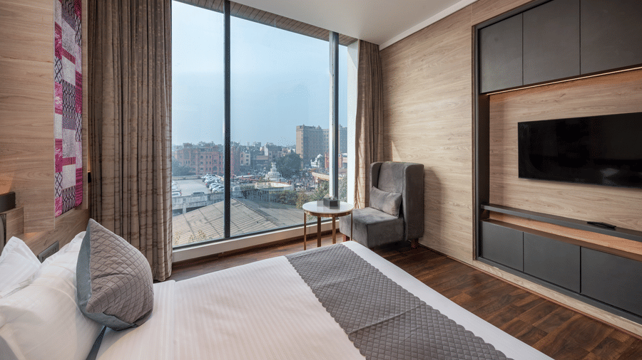 A bedroom with a king size bed, chair, table, wall-mounted television, and large window, at DLS Hotels Amritsar.