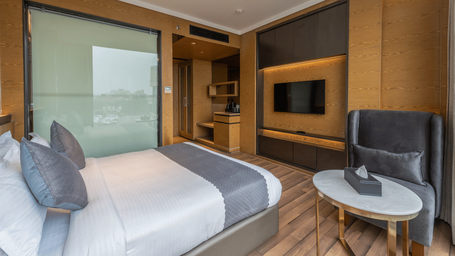 A bedroom with a king size bed, armchair, table, glass partition, wall-mounted TV, and wooden flooring at DLS Hotels Amritsar.