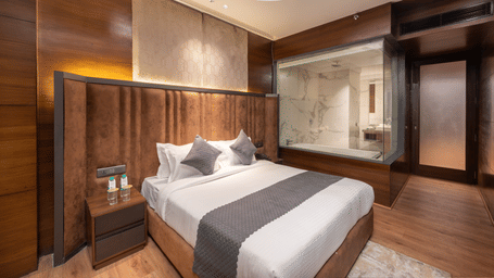 A room with a king size bed, side table, ceiling fan, and a glass partition leading to another area at DLS Hotels Amritsar.