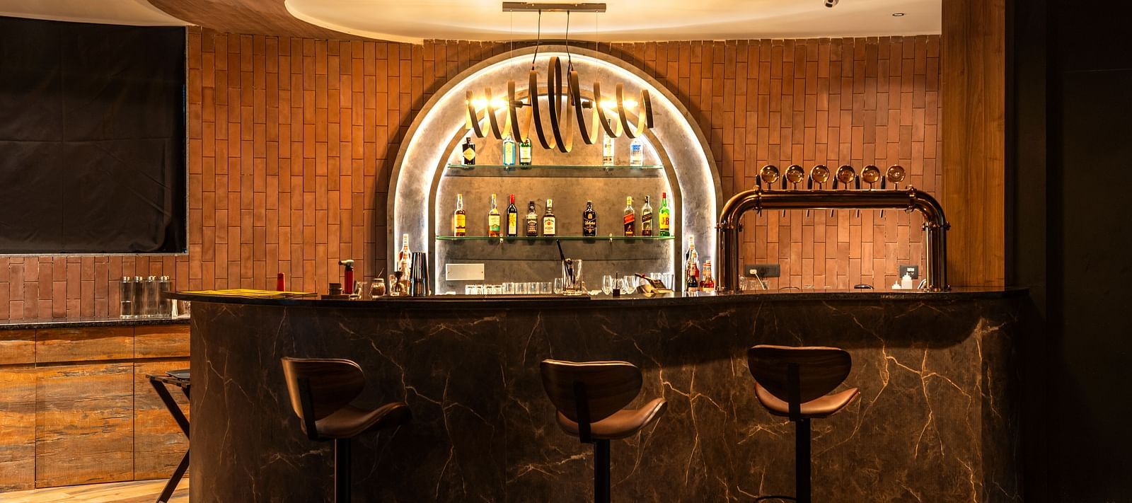 Rhythm & Brews equipped with high chairs and a variety of bottles displayed on shelves, illuminated by warm lighting at The Qualia Resort Club & Brewery, Udaipur.