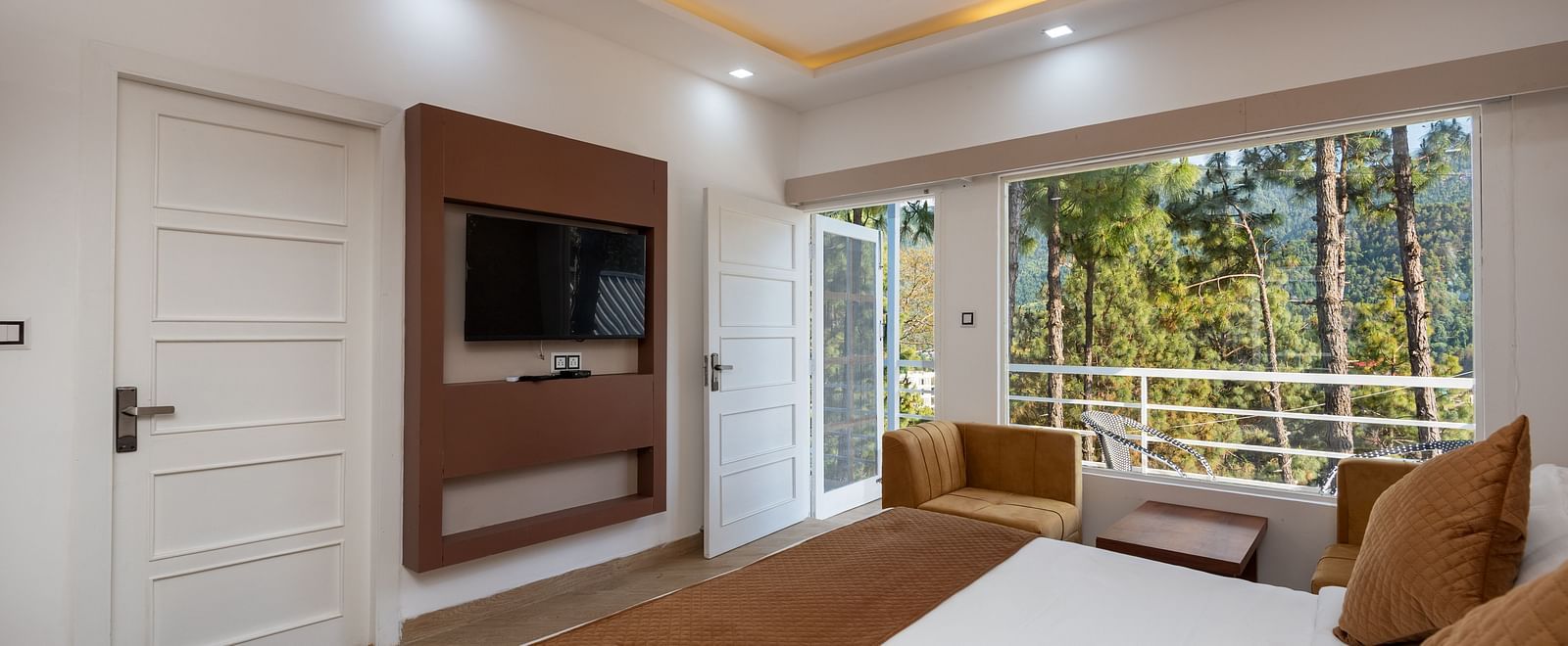 Modern hotel room with a double bed featuring brown accents, a wall-mounted flat-screen television, comfortable armchairs, and large windows overlooking a forested view - DLS Nature Trinket Resort, Dalhousie.