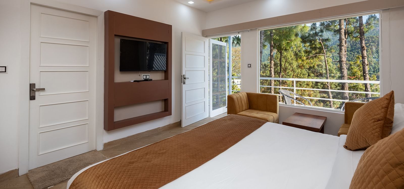 Modern hotel room with a double bed featuring brown accents, a wall-mounted flat-screen television, comfortable armchairs, and large windows overlooking a forested view - DLS Nature Trinket Resort, Dalhousie.