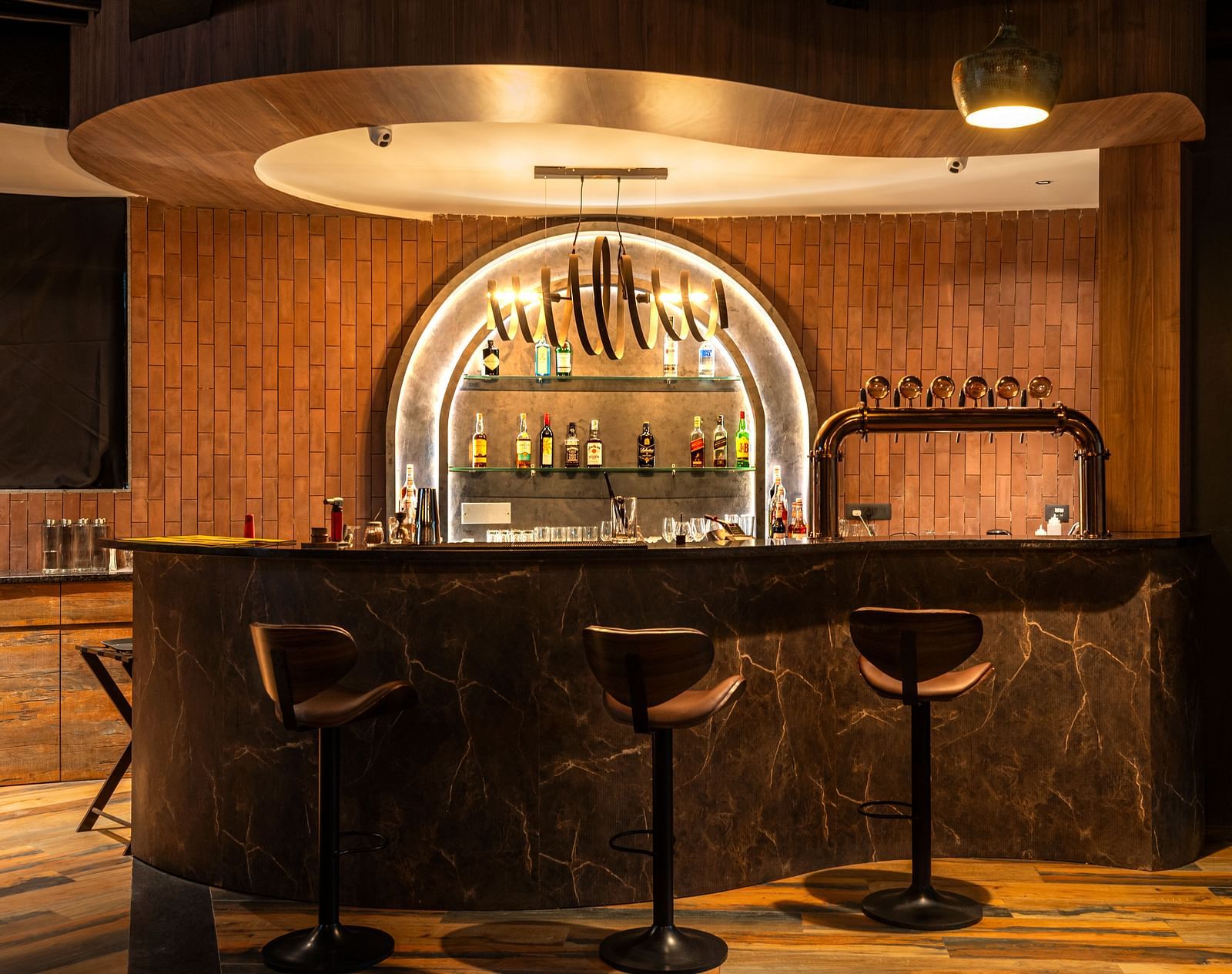 Rhythm & Brews equipped with high chairs and a variety of bottles displayed on shelves, illuminated by warm lighting at The Qualia Resort Club & Brewery, Udaipur.
