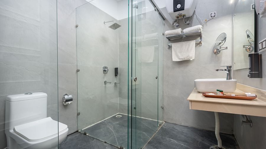 A contemporary bathroom with a glass-enclosed shower area, white toilet, wall-mounted water heater, and a sleek wooden countertop basin, all set against light grey tiled walls and a dark slate floor - DLS Nature Trinket Resort, Dalhousie.