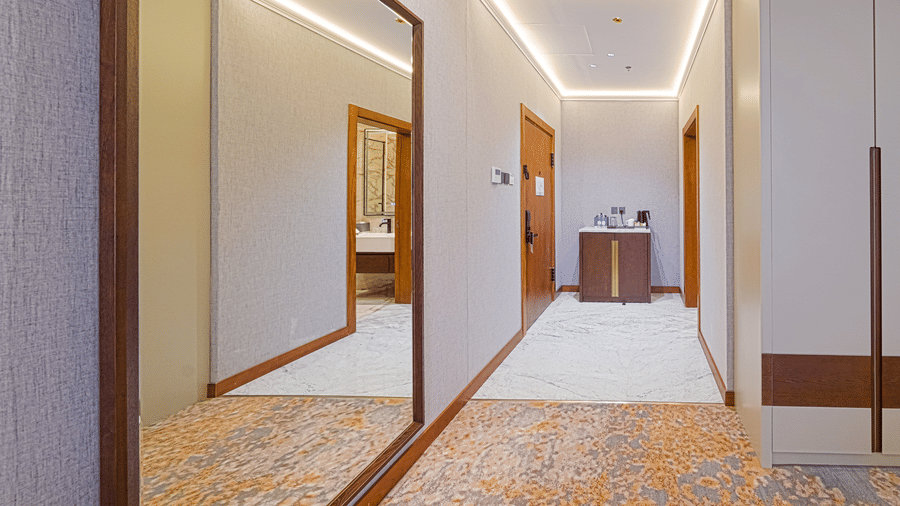 Doorway of a Deluxe Twin Room at Grand Iva Hotel, Abha, showcasing a vanity table, a wall-mounted full length mirror, overhead lighting, with the doorway leading to a bedroom with twin beds and upholstered seating area.