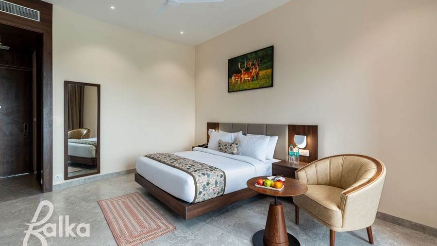 A clean bedroom at Osian Valley Resort & Spa featuring a double bed with a patterned runner, a side table with fruit, and a full-length mirror next to a dark wooden door.
