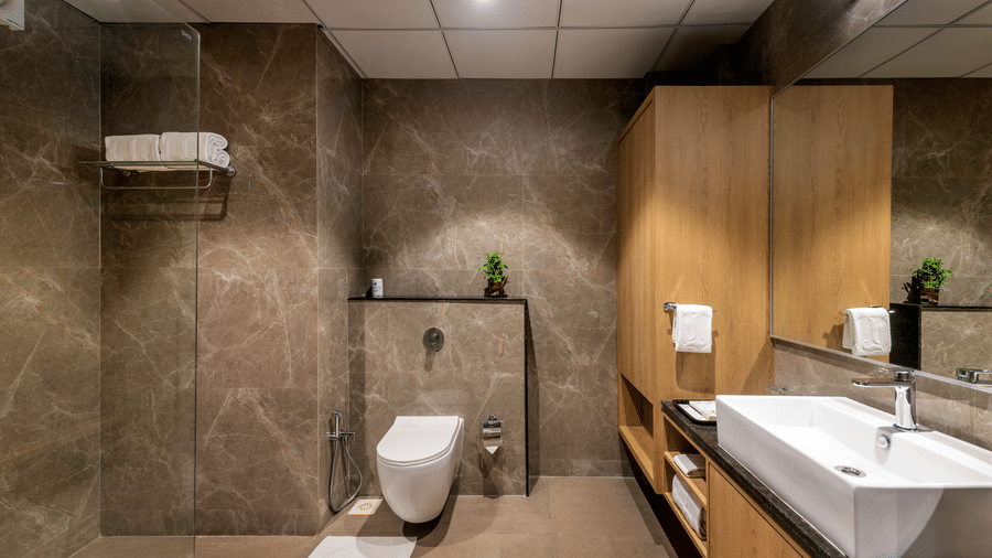 A modern bathroom in Lords Somnath, with sleek marble walls, featuring a walk-in shower, wall-mounted toilet, large mirror, and counter top basin. 