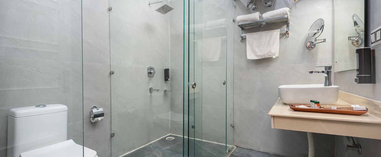 A contemporary bathroom with a glass-enclosed shower area, white toilet, wall-mounted water heater, and a sleek wooden countertop basin, all set against light grey tiled walls and a dark slate floor - DLS Nature Trinket Resort, Dalhousie.