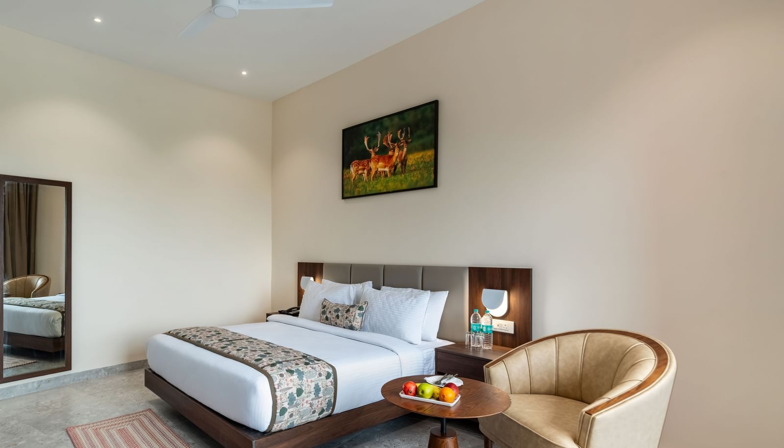 A clean bedroom at Osian Valley Resort & Spa featuring a double bed with a patterned runner, a side table with fruit, and a full-length mirror next to a dark wooden door.