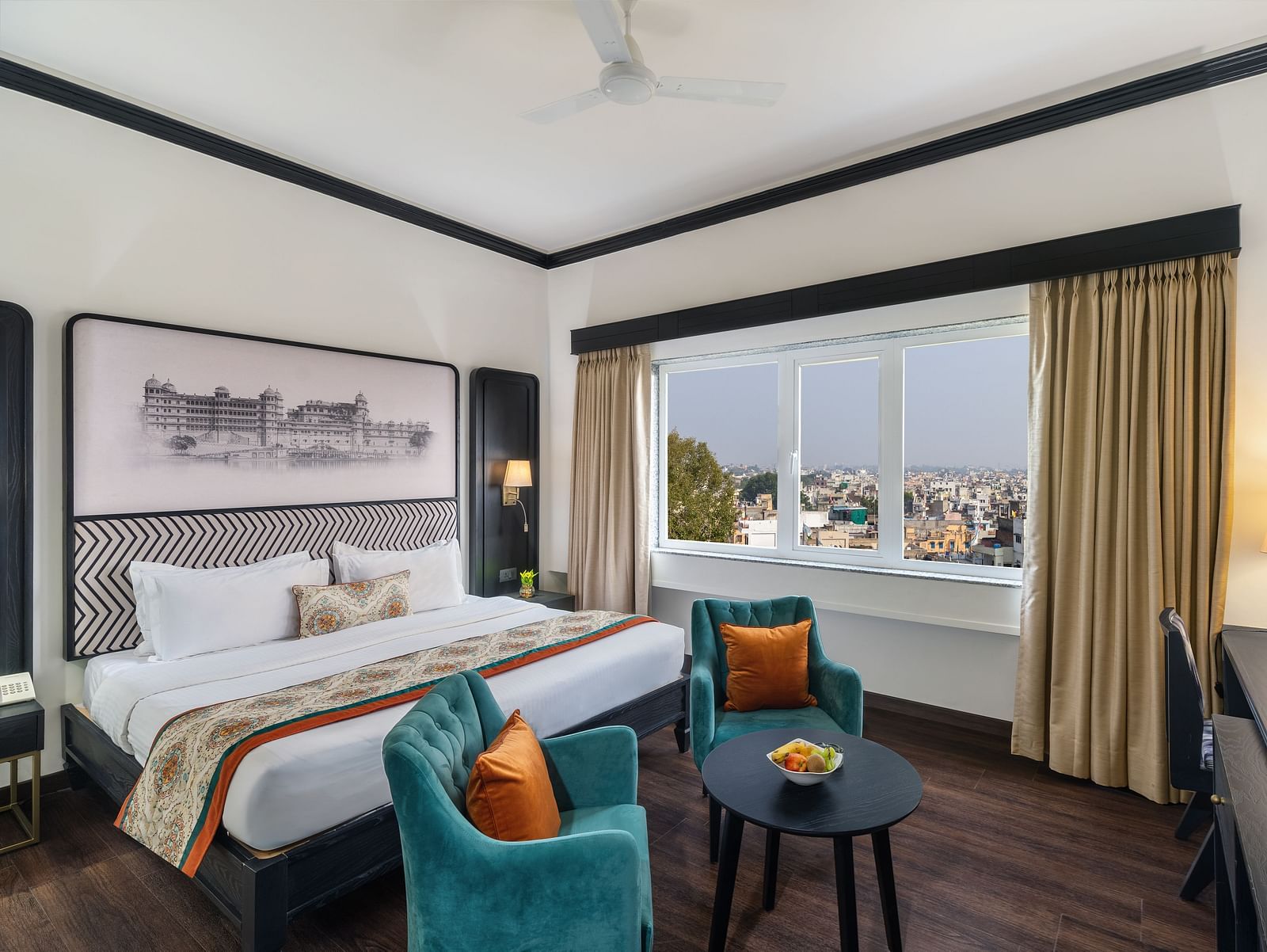a spacious room featuring a king size bed, two comfortable green chairs, a cabinet with a table lamp and a window overlooking the city - Hotel Rajdarshan, Udaipur.