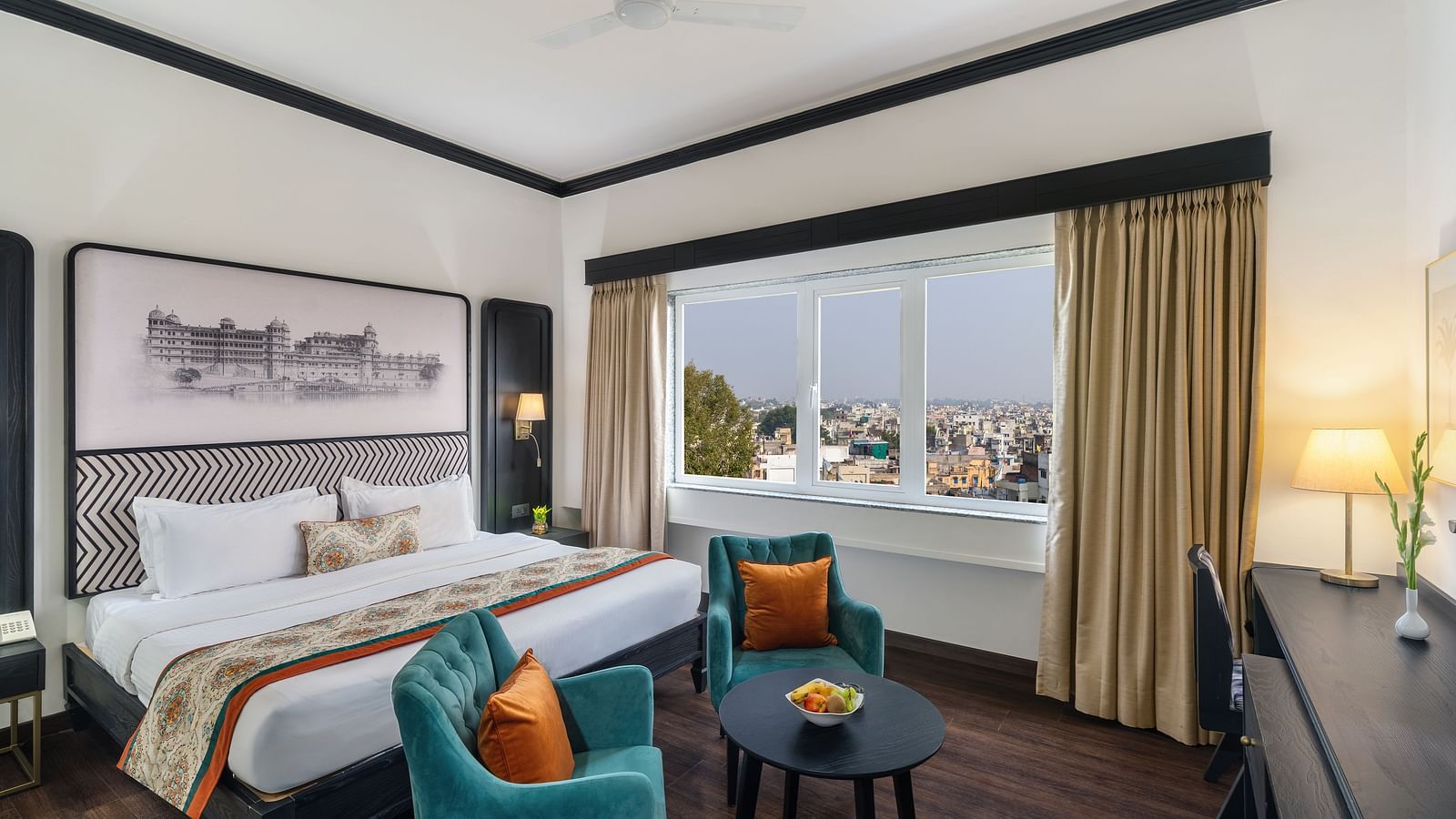 a spacious room featuring a king size bed, two comfortable green chairs, a cabinet with a table lamp and a window overlooking the city - Hotel Rajdarshan, Udaipur.