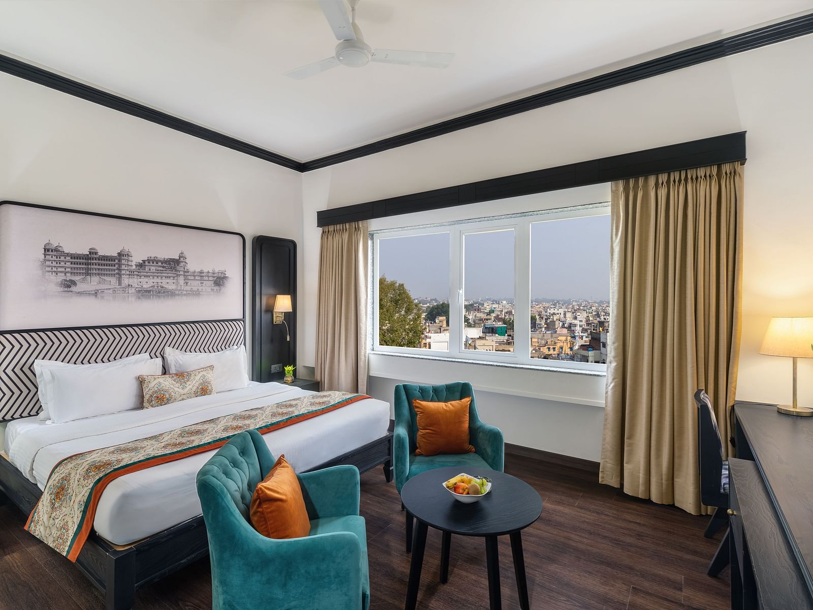 a spacious room featuring a king size bed, two comfortable green chairs, a cabinet with a table lamp and a window overlooking the city - Hotel Rajdarshan, Udaipur.
