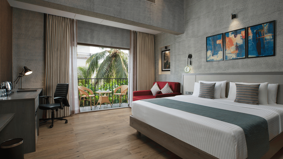 Bright hotel room at Ocean Palms Goa with a king bed, work desk, large balcony doors overlooking tropical palm trees, wooden flooring, and contemporary decor.