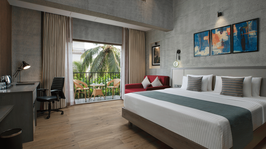 Bright hotel room at Ocean Palms Goa with a king bed, work desk, large balcony doors overlooking tropical palm trees, wooden flooring, and contemporary decor.