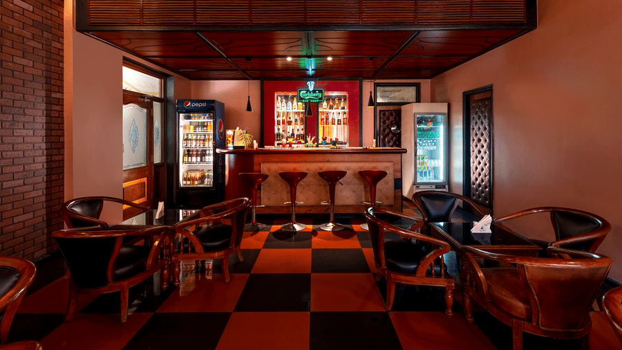 An indoor bar area features a wooden counter, display refrigerators, tables, and wooden chairs at Amantra Shilpi Resort, Udaipur.