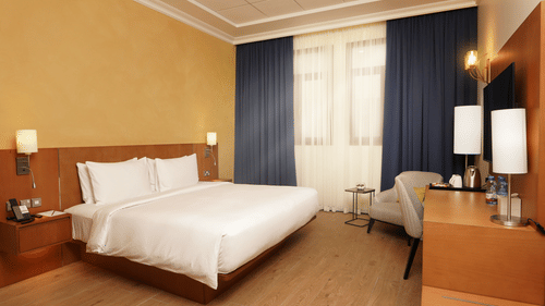 A corner view of two bedroom apartment at La Maison, Doha, with a made bed, a curtained window, and a small table.
