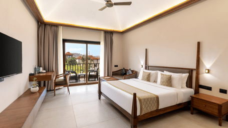 A bright hotel room features a large wooden bed and glass doors leading out to a sunny outdoor balcony - Sajjan Bagh Resort and Spa, Kumbhalgarh.