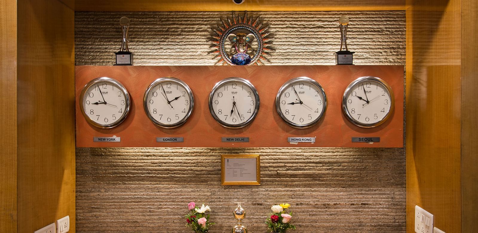 A display of clocks showing different time zones, mounted on a wooden panel - Lords Eco Inn Dahej