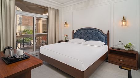A spacious bed with a cushioned headboard, flanked by bedside tables on each side, and large windows with curtains pulled to the side | Hotel Ganga Maiya