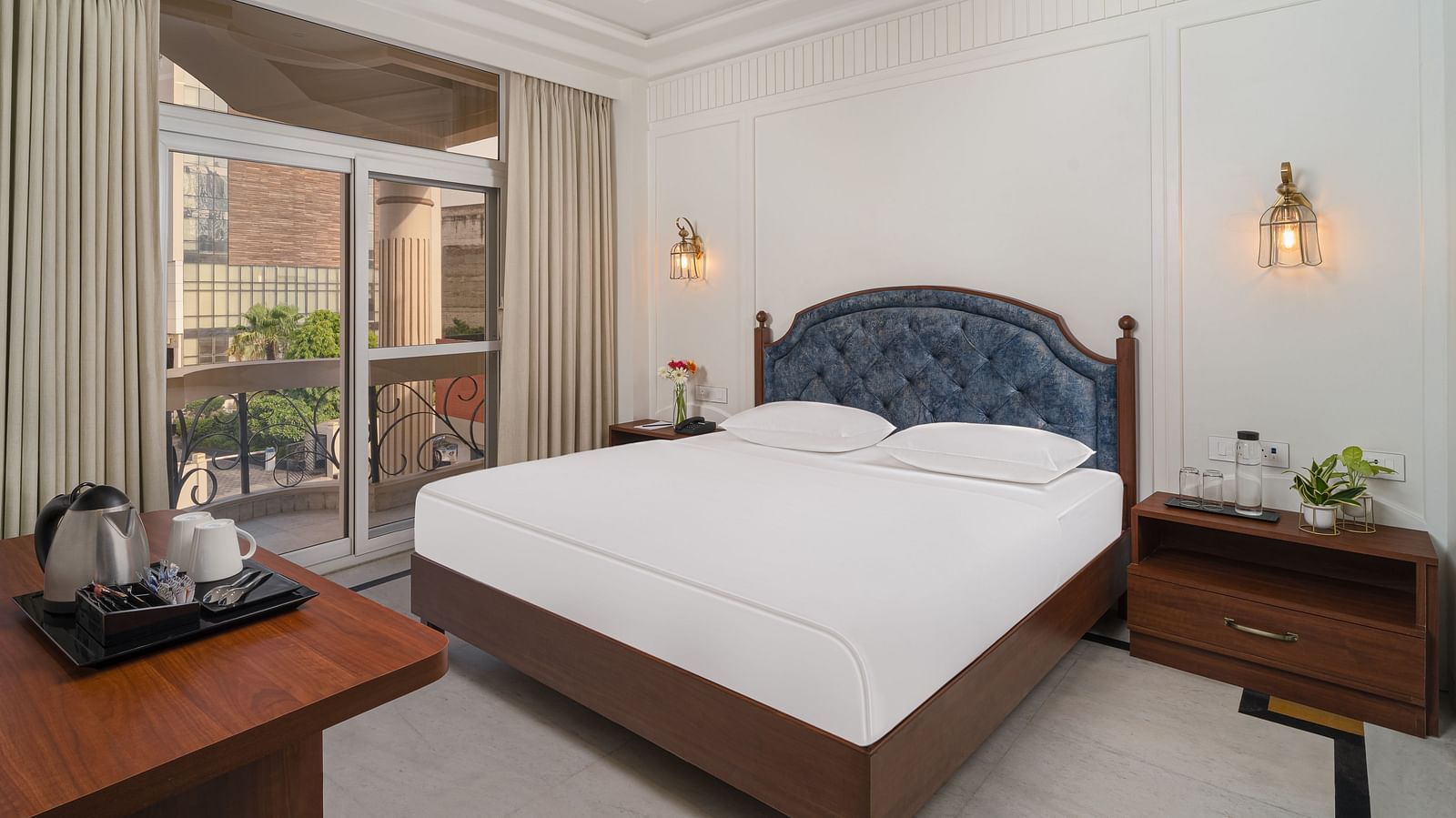 A spacious bed with a cushioned headboard, flanked by bedside tables on each side, and large windows with curtains pulled to the side | Hotel Ganga Maiya