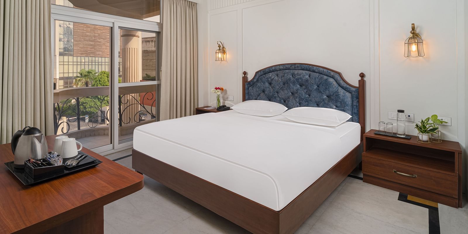 A spacious bed with a cushioned headboard, flanked by bedside tables on each side, and large windows with curtains pulled to the side | Hotel Ganga Maiya