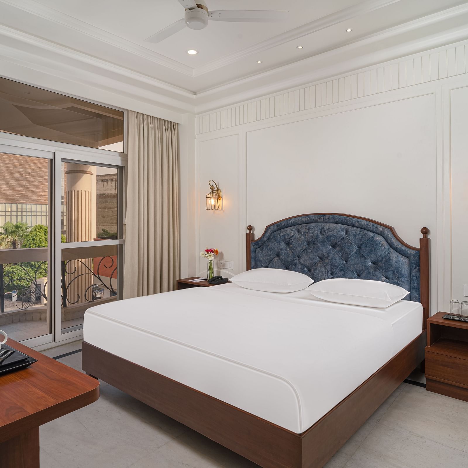 A spacious bed with a cushioned headboard, flanked by bedside tables on each side, and large windows with curtains pulled to the side | Hotel Ganga Maiya
