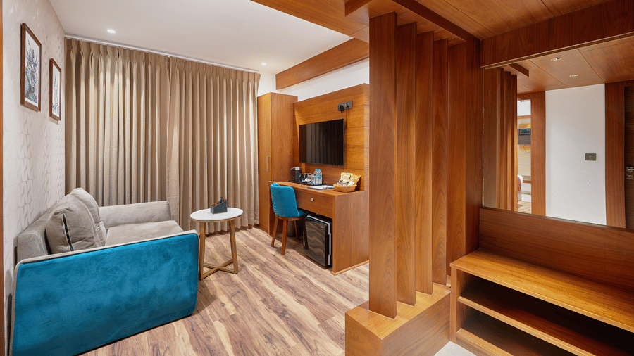 View of a suite living area at QWhite Crescent Luxury Hotel by Dimora Kochi with a teal sofa, wooden partition, work desk, and warm flooring.