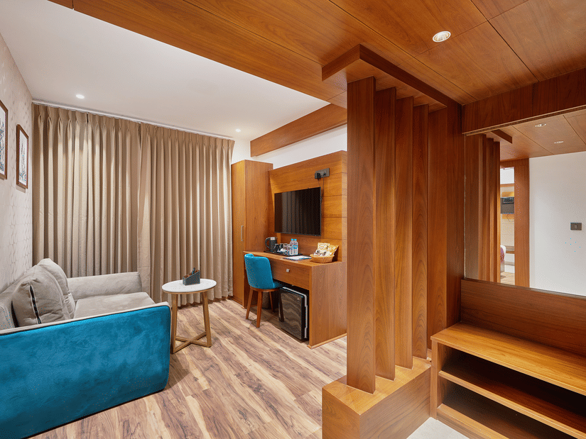 View of a suite living area at QWhite Crescent Luxury Hotel by Dimora Kochi with a teal sofa, wooden partition, work desk, and warm flooring.