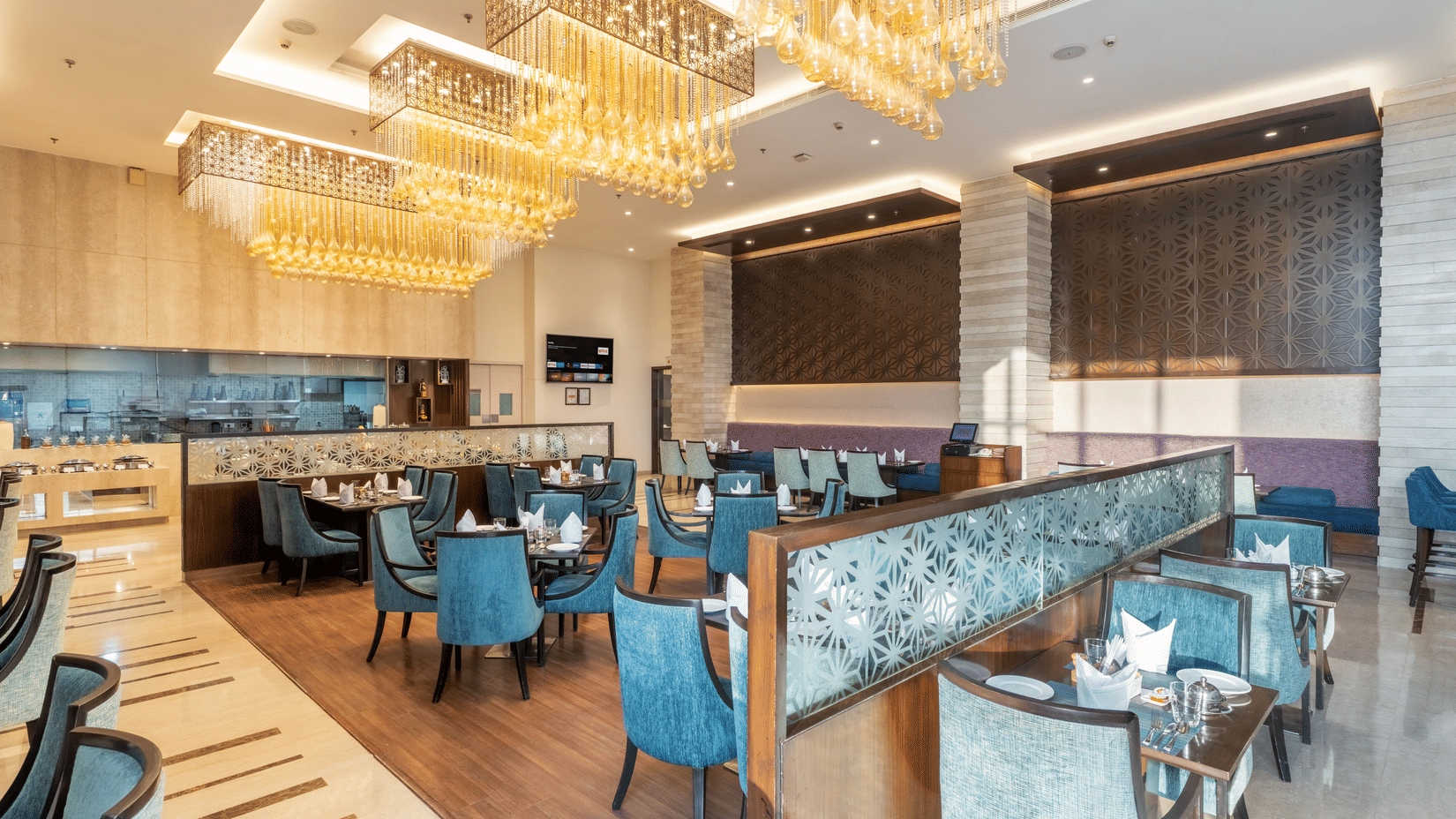 A dining section at Nirvana Sarovar Portico, Gorakhpur featuring padded chairs, wooden tables, and overhead chandeliers. 