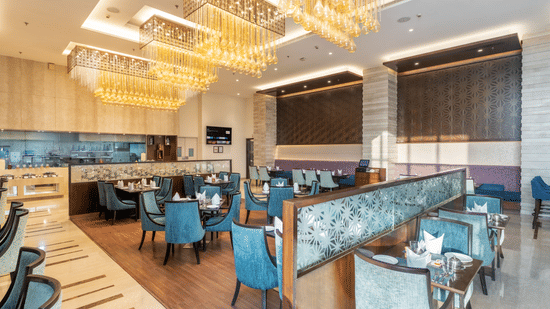 An indoor dining space at Nirvana Sarovar Portico, Gorakhpur featuring dining tables, padded chairs, and large light fixtures.