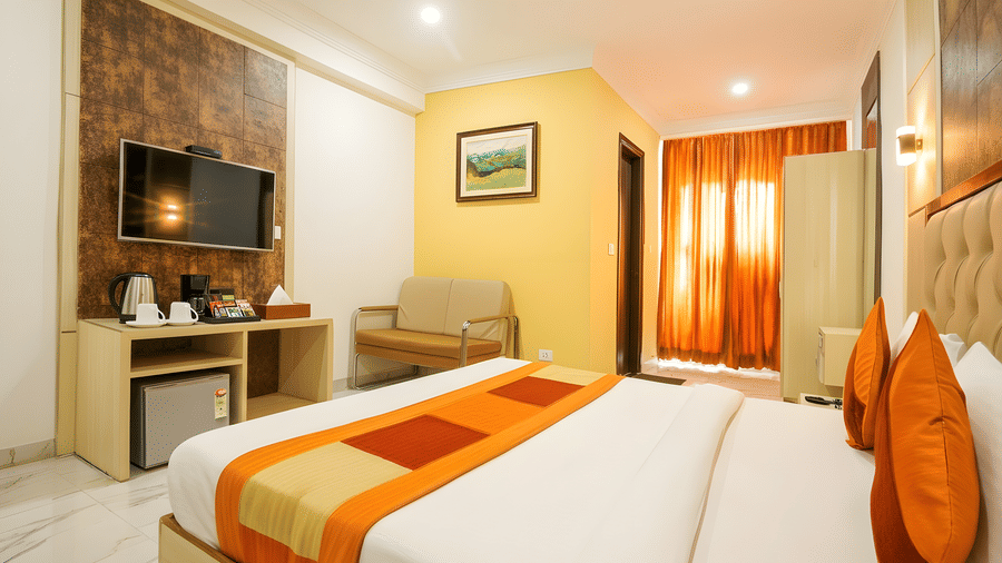 A brightly lit room at Hotel Apple Park Delhi featuring a bed with orange and brown runners, orange curtains, and a mini-fridge housed in a wooden unit.