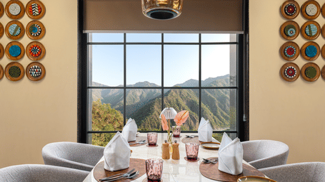 Indoor dining table with placemats and napkins set beside a large grid window overlooking mountain scenery.