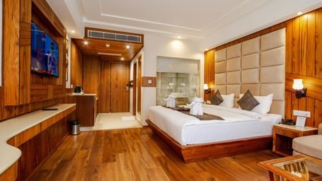 Spacious hotel room with modern furnishings, a large comfortable bed, wall-mounted TV, wooden flooring, and a glass-enclosed bathroom area.